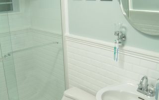 transitional guest hallway bathroom with white subway tile wainscot, light blue walls, pedestal ADA sink , tilt mirror, chrome fixtures, glass shower enclosure, and layered lighting plan