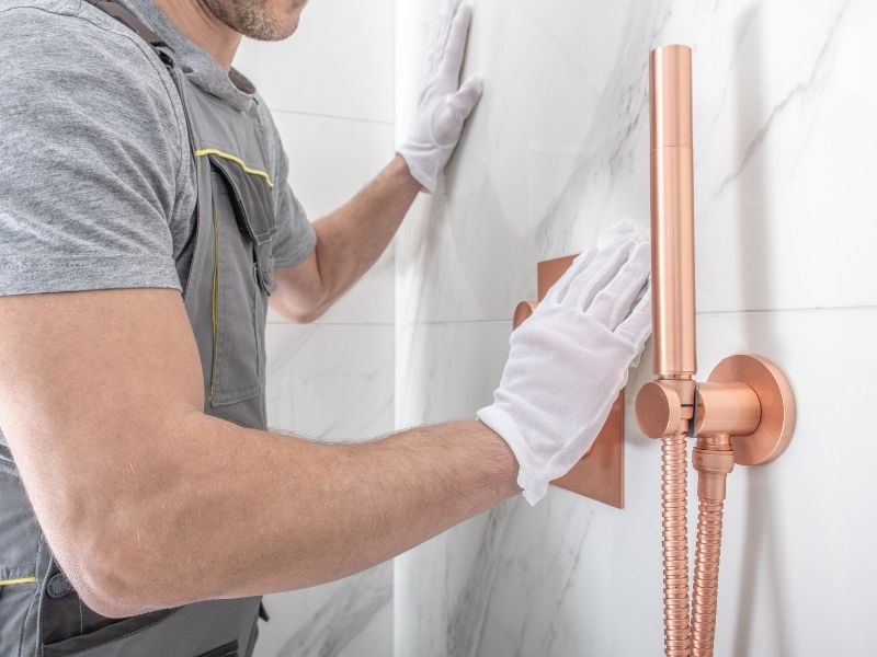 The 13 Essential Signs that Your Bathroom Needs a Remodel - photo of a plumber fitting copper fixtures to a marble shower wall.